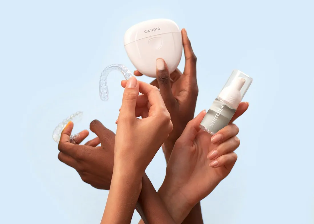 Hands holding a clear orthodontic aligner, storage case and whitening pen against a pale blue background
