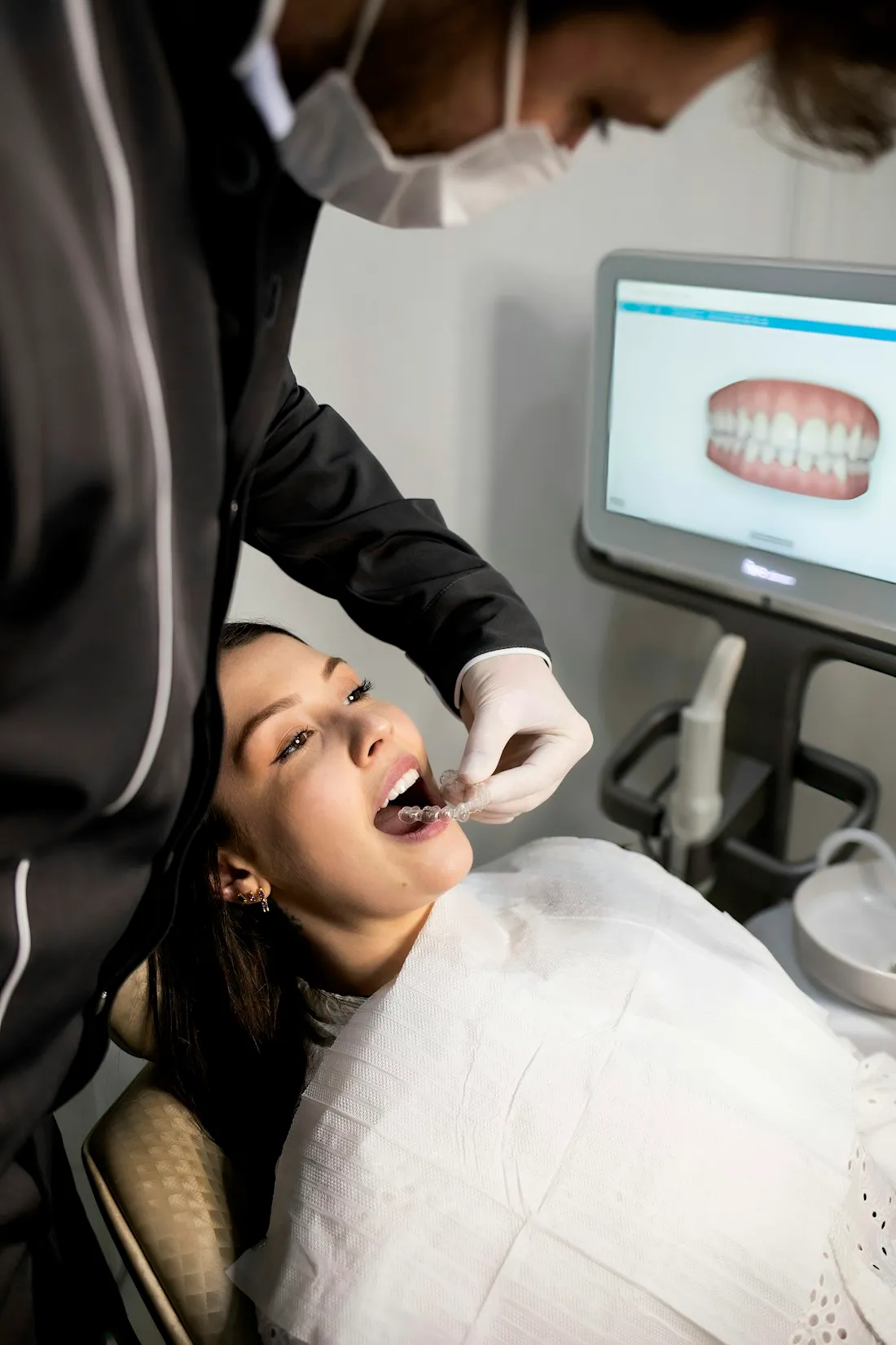 Dentist fitting a clear aligner for a patient while a digital scan of her teeth is displayed on the monitor behind