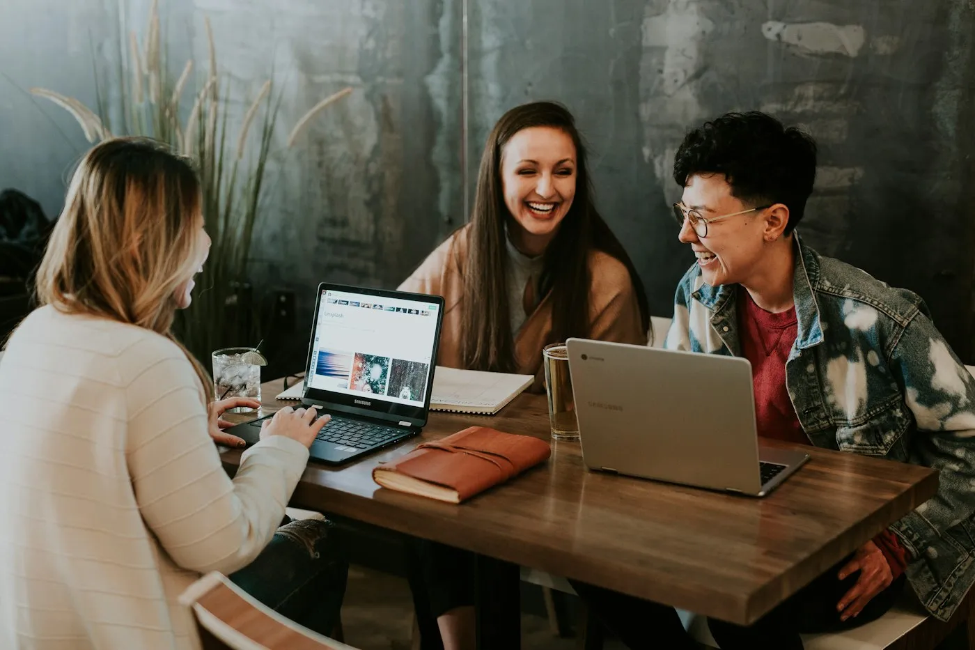 Three colleagues laughing while working on laptops at a cafe table