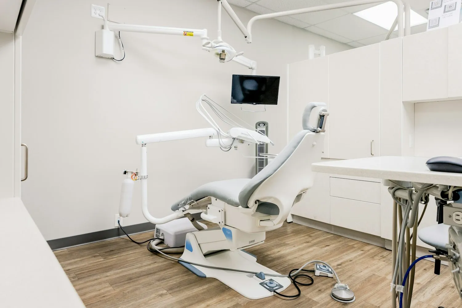 Clean modern dental operatory with white cabinetry, a wooden floor and a treatment chair under daylight